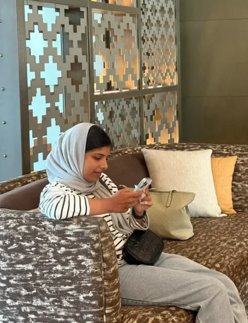 A person sits on a patterned sofa, looking at a smartphone, with a handbag beside them and decorative geometric screens in the background.